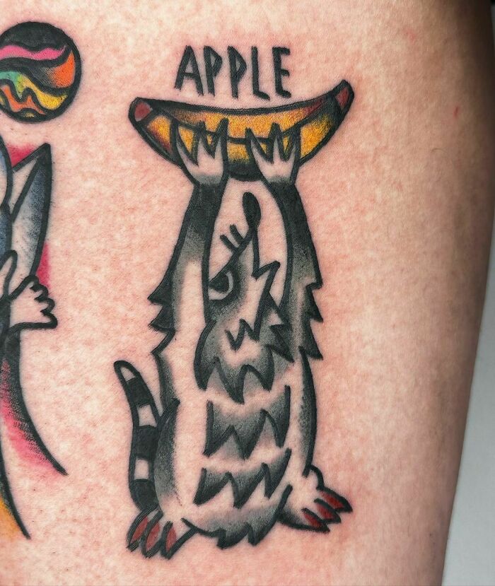 A creative animal tattoo with a raccoon holding a banana labeled "Apple."