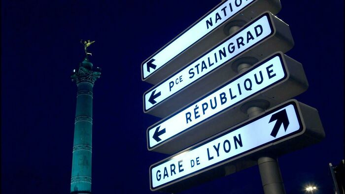 Street signs in a city at night with a column monument in the background.