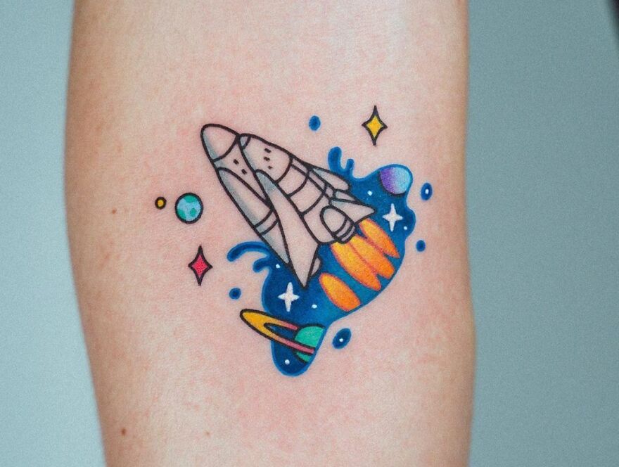 Space tattoo featuring a colorful rocket blasting off with stars and planets in the background.