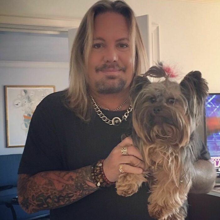 Celebrity holding a small dog, wearing a black shirt and jewelry, standing indoors.