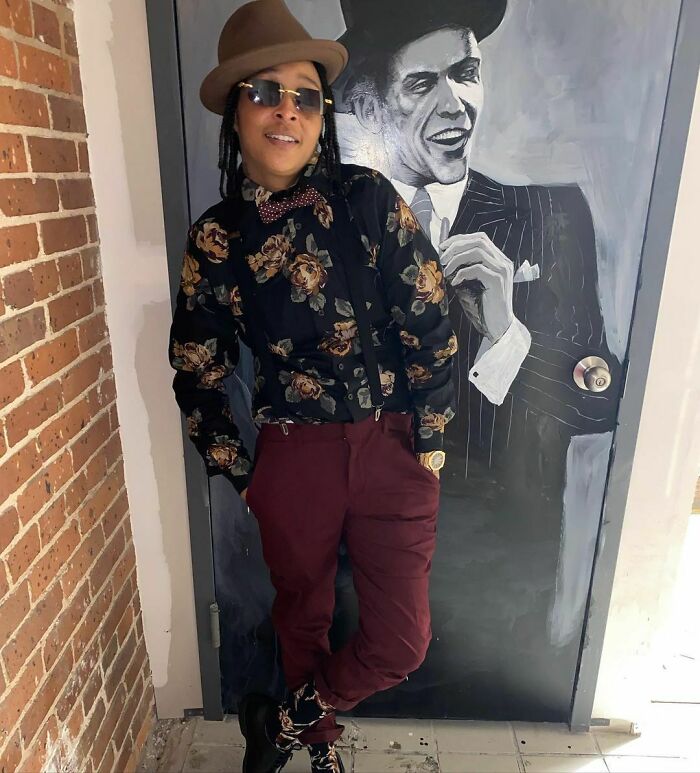 Person in floral shirt and hat posing next to a painted mural, reflecting celebrity style and vibe.