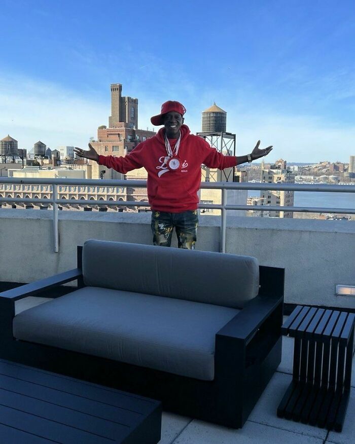 Celebrity in a red hoodie poses on a rooftop with cityscape in the background.