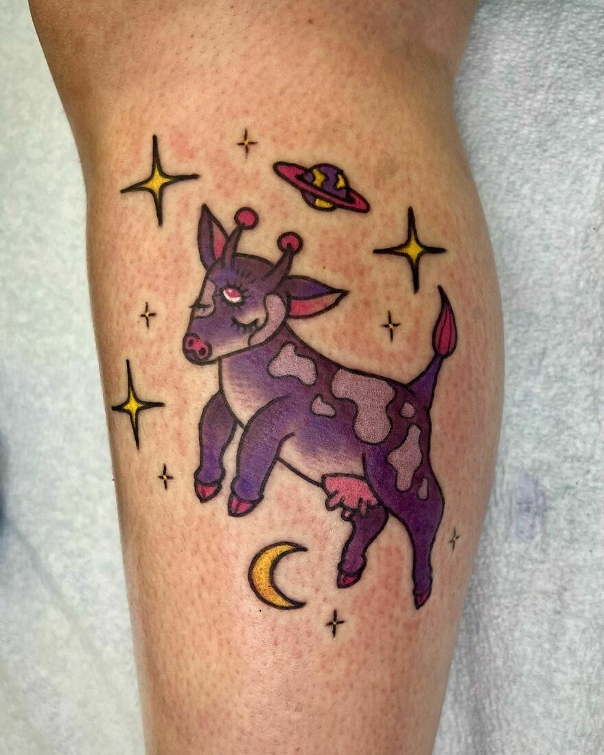 A cosmic-themed tattoo features a purple cow with antennae, a UFO, stars, and a crescent moon, representing outer space cool.