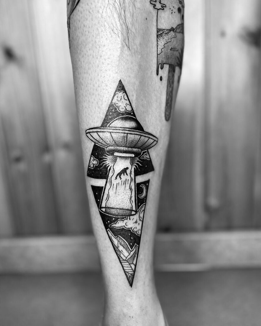 Black and white space tattoo of a UFO abducting a wolf, with mountains and clouds, showcasing outer space art on a leg.