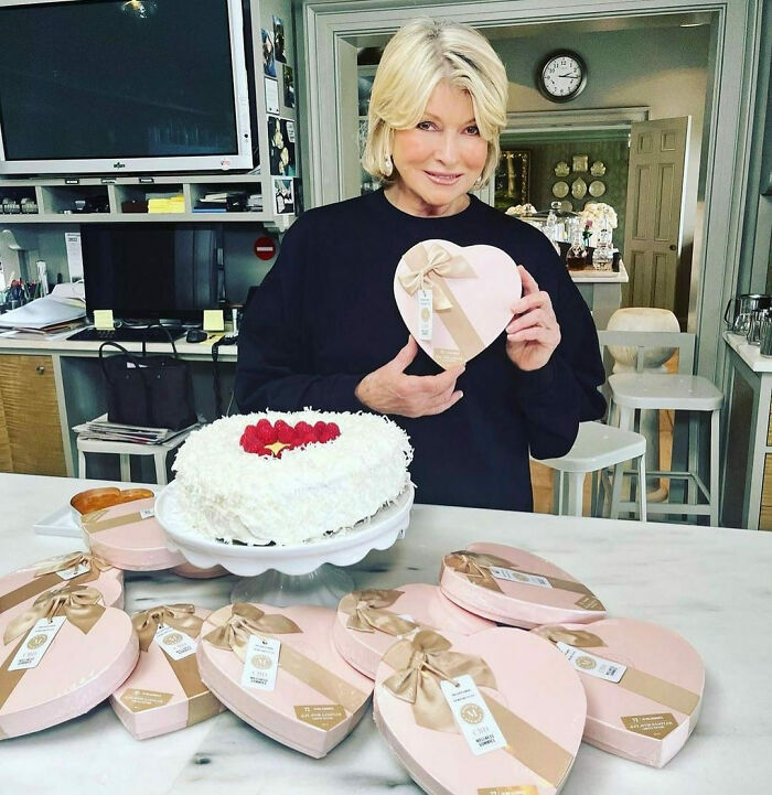 Celebrity holding heart-shaped box near cake in kitchen, surrounded by similar boxes.