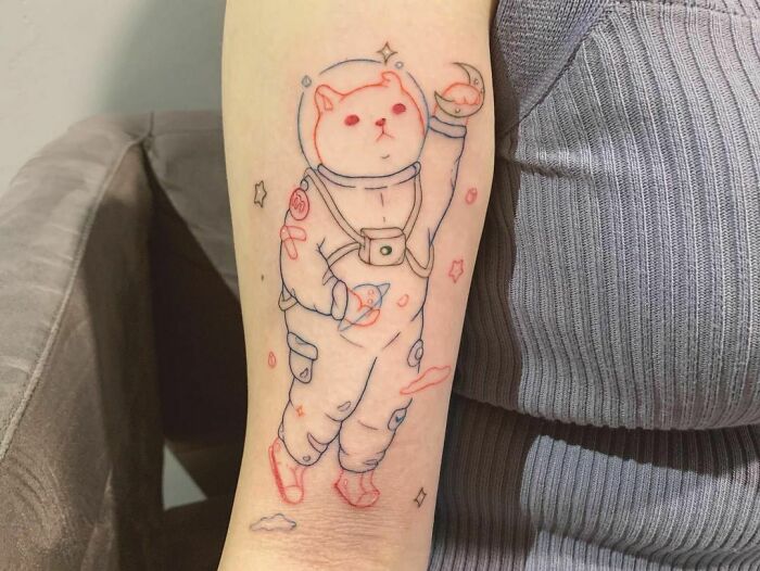 Cute space tattoo of a cat in an astronaut suit on someone's arm, featuring stars and planets.