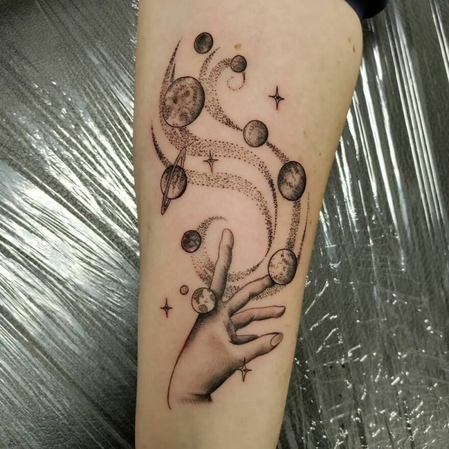 Space tattoo featuring a hand reaching for planets and stars in a cosmic design on forearm.