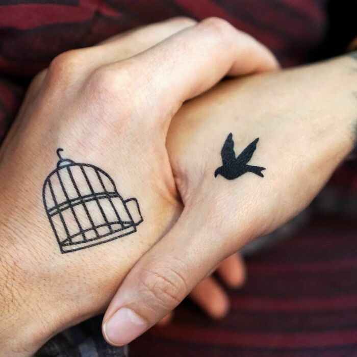 Matching tattoos of a birdcage and bird on hands, symbolizing freedom and connection.