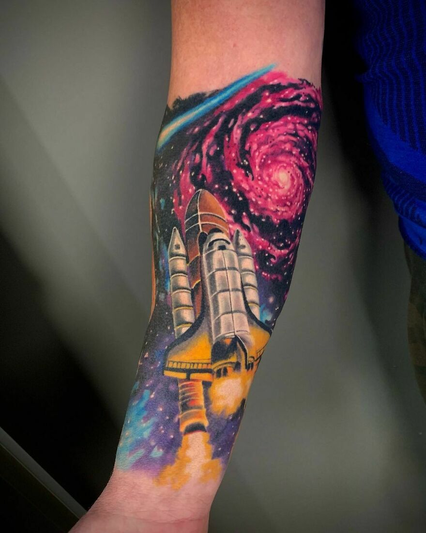 Space tattoo featuring a detailed rocket launching with a colorful spiral galaxy background on a forearm.