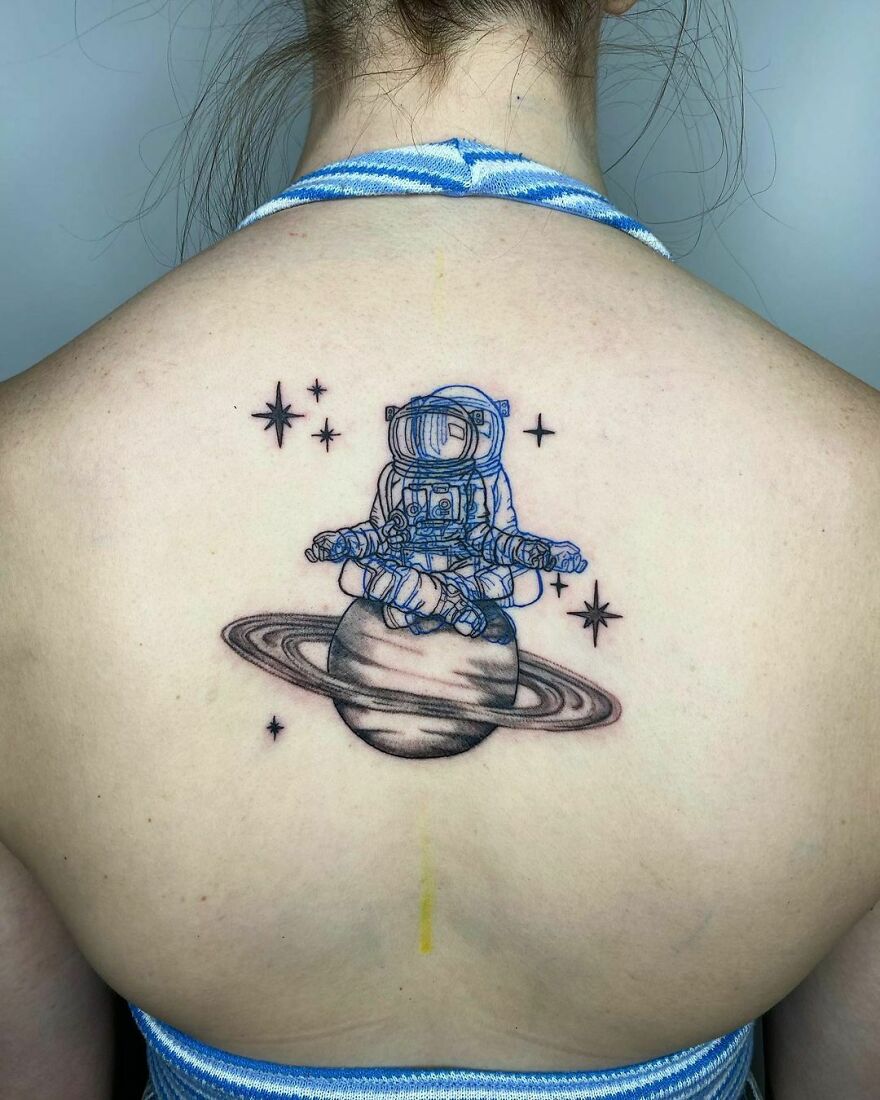 Space tattoo of an astronaut meditating on a planet with rings, surrounded by stars, on a person's back.