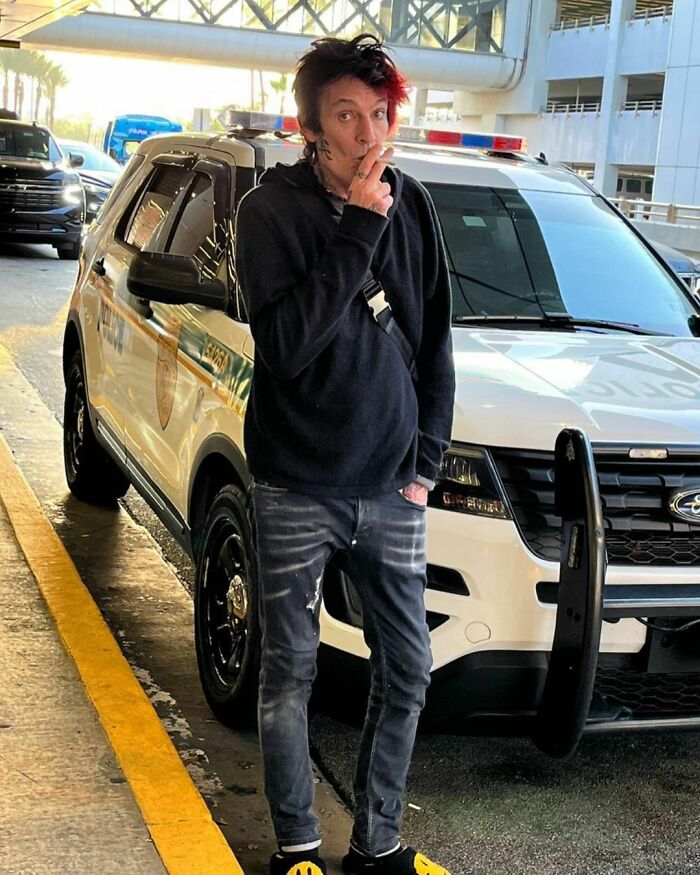 Celebrity standing by a police car, wearing dark clothing and yellow shoes, possibly linked to minor or severe crimes.