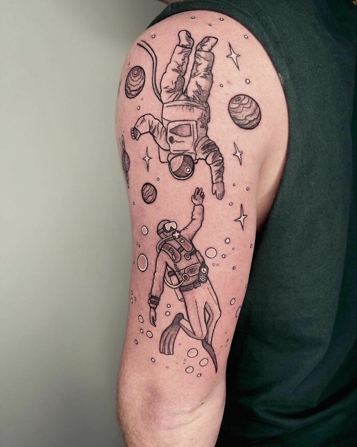 Space-themed tattoo with astronauts and planets on an upper arm.