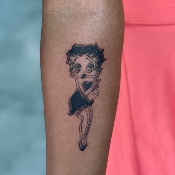 Tattoo of a cartoon character on an arm, with a pink dress in the background.