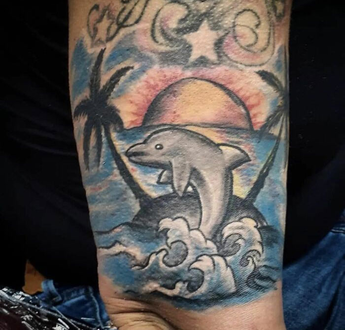 A poorly designed tattoo of a dolphin jumping out of waves with a sunset and palm trees in the background.