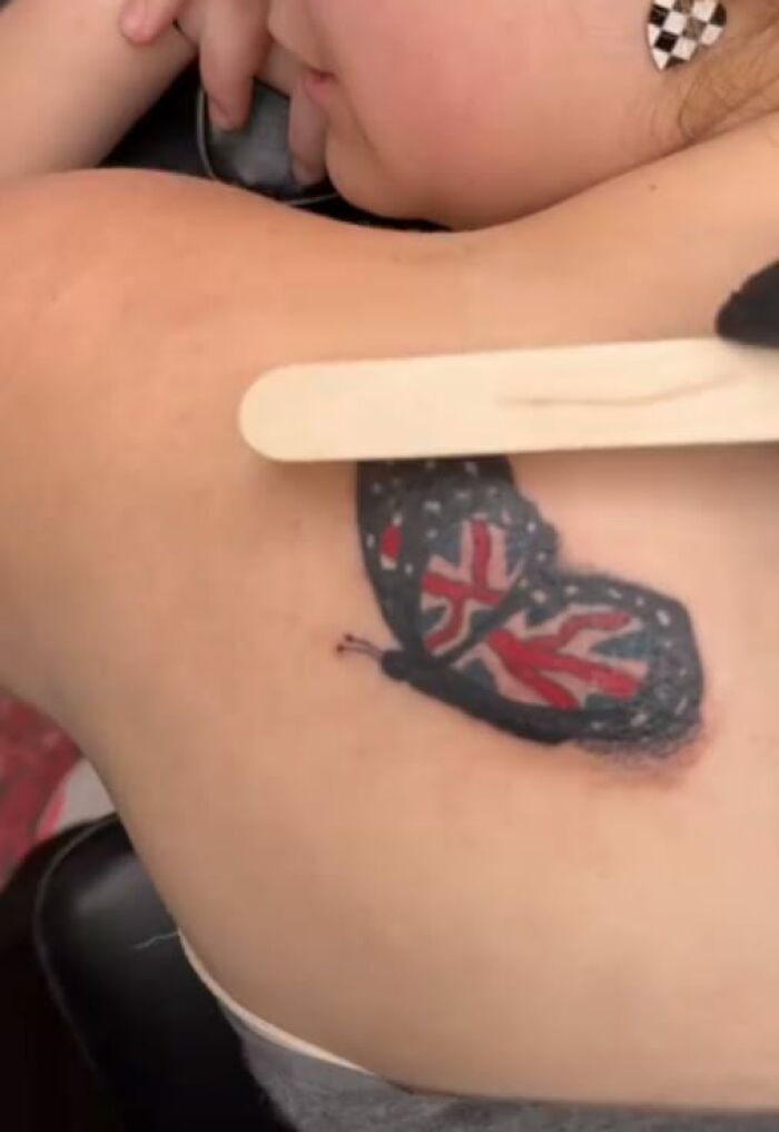 Person with a poorly drawn butterfly tattoo featuring a flag design on their shoulder.