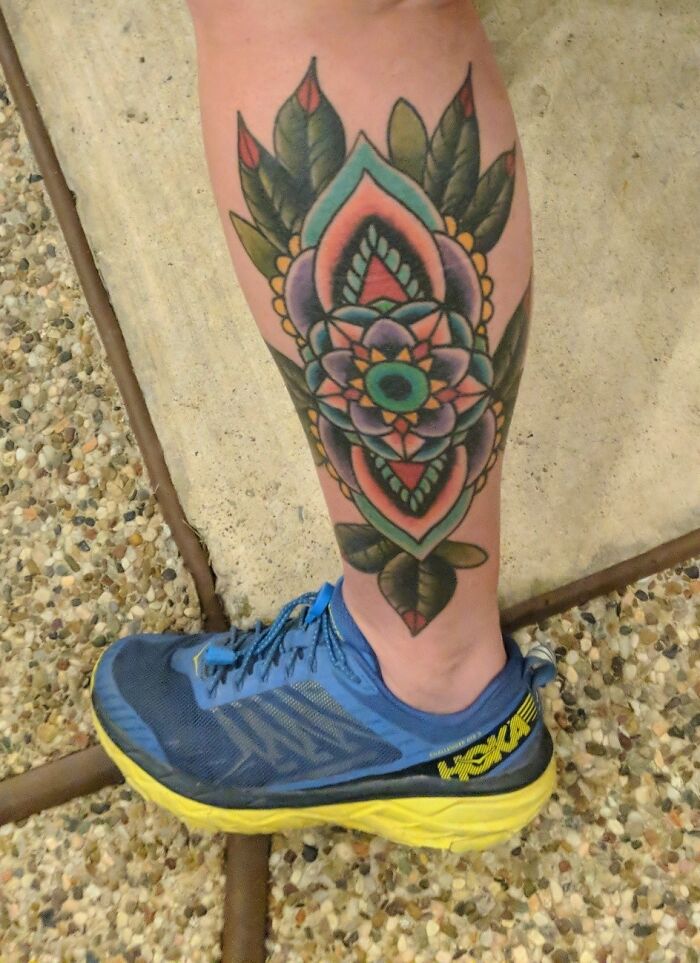 Colorful tattoo on leg with a floral design next to blue sneakers on a pebbled surface.
