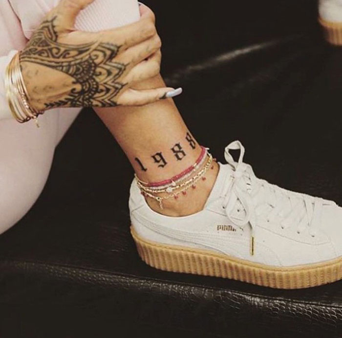 Celebrity tattoo on ankle reading "1988," with ornate hand tattoo and white sneakers.
