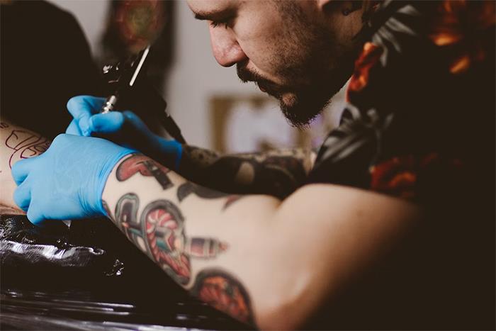 Tattoo artist inking a client's arm, showing detailed tattoos and wearing blue gloves.