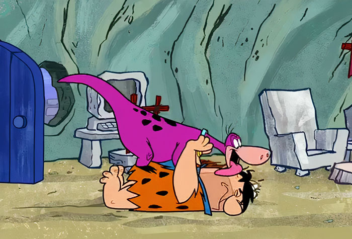 Cartoon dog and caveman playing on the floor in a prehistoric home setting.