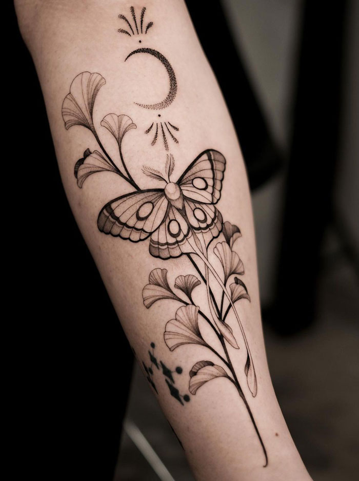 Arm with a flower tattoo featuring a butterfly and crescent moon, showcasing intricate design blossoming on the skin.