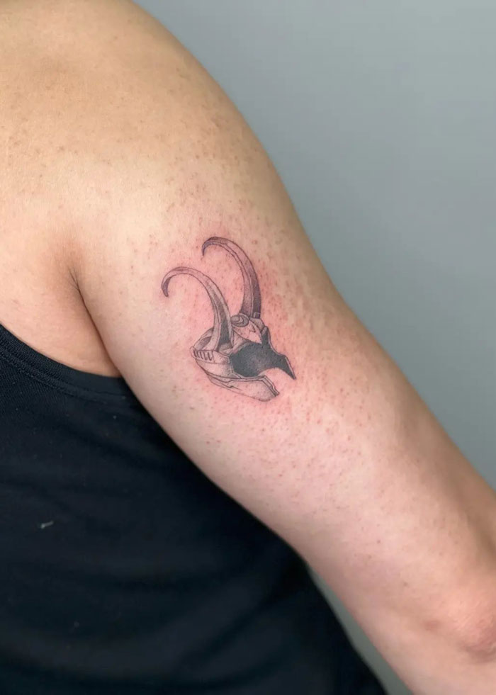 Marvel tattoo of Loki helmet on upper arm in black ink, showcasing intricate design and superhero theme.