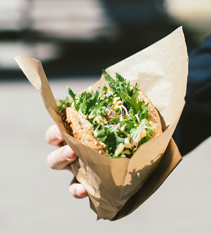person holding brown paper bag with vegetable sandwich