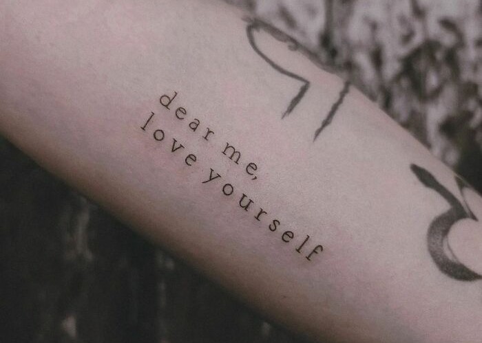 Tattoo on arm with text "dear me, love yourself," embodying self-love and empowerment.