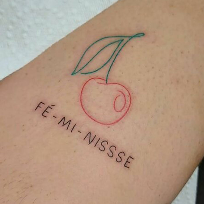 Cherry tattoo with "Fé-mi-nisse" text promotes self-love on skin.