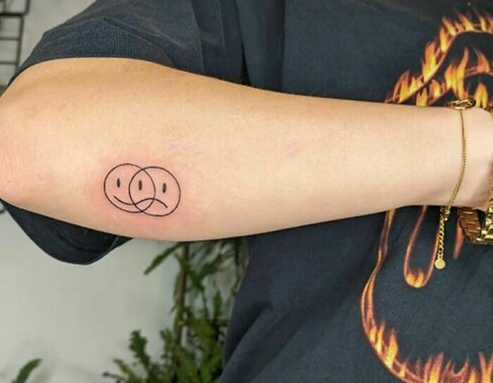 Arm with minimalist self-love tattoo showing overlapping happy and sad faces.