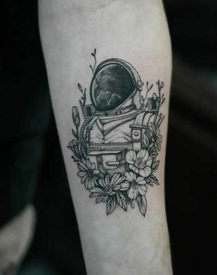 Space-themed tattoo featuring an astronaut helmet surrounded by flowers on a forearm.