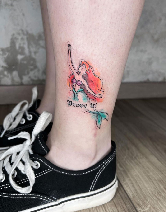 Watercolor tattoo of a mermaid with "Prove it!" text on a person's leg, near black sneakers.