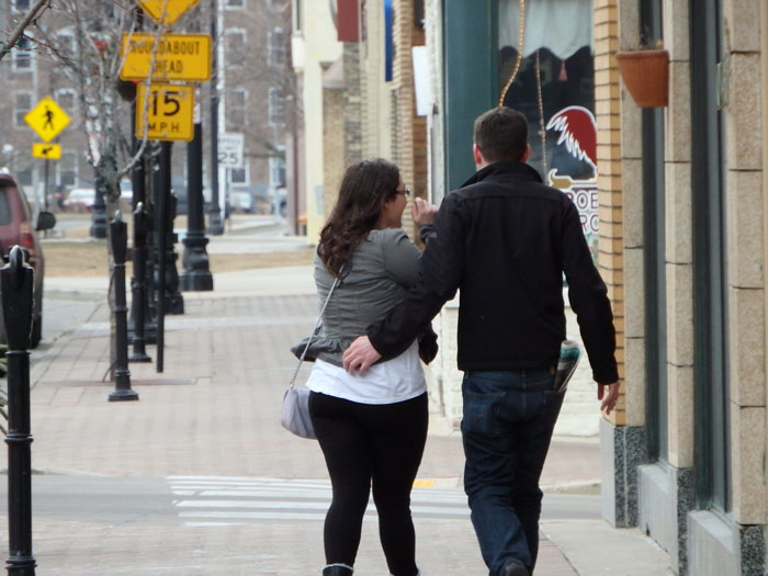 Couple walking down a city street, with storefronts and a pedestrian crossing sign in the background.
