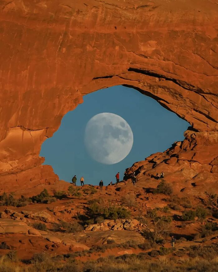 Moon visible through rock arch, with people exploring below; an amusing photo from popular Instagram page.