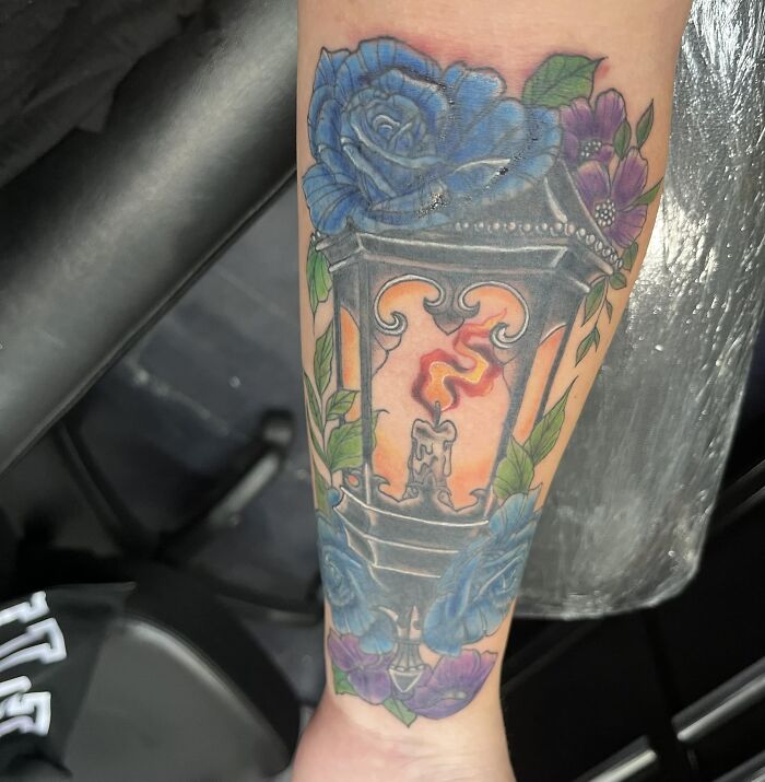 Colorful lantern tattoo with blue roses and greenery on forearm.