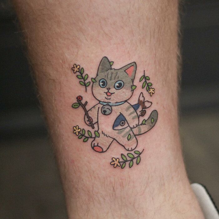 Adorable animal-themed tattoo of a cartoon cat holding a twig, surrounded by flowers.