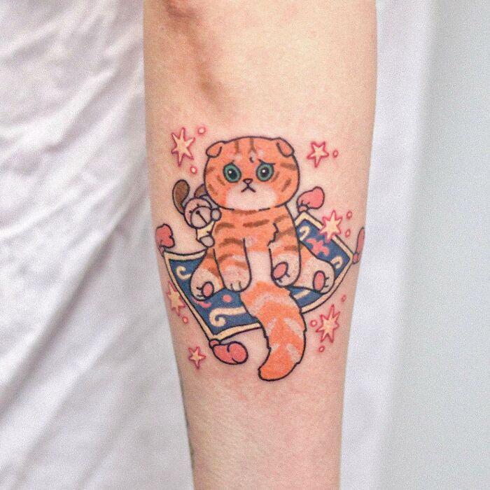 Adorable animal-themed tattoo of a cartoon cat on a magic carpet with stars, designed by a talented artist.