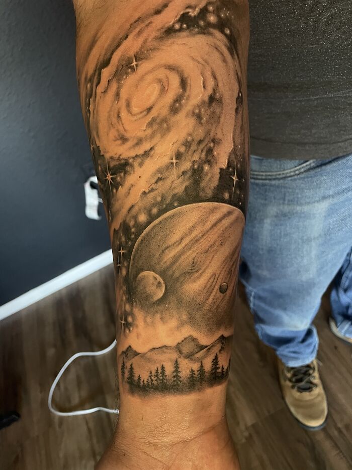 Arm tattoo featuring a galaxy scene with planets and forest, showcasing cool tattoo design.