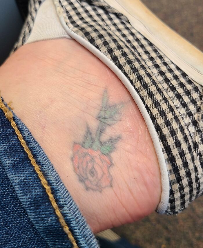 Aged tattoo of a rose on ankle, partially faded, showing ink changes over the years, with checkered shoe and denim visible.