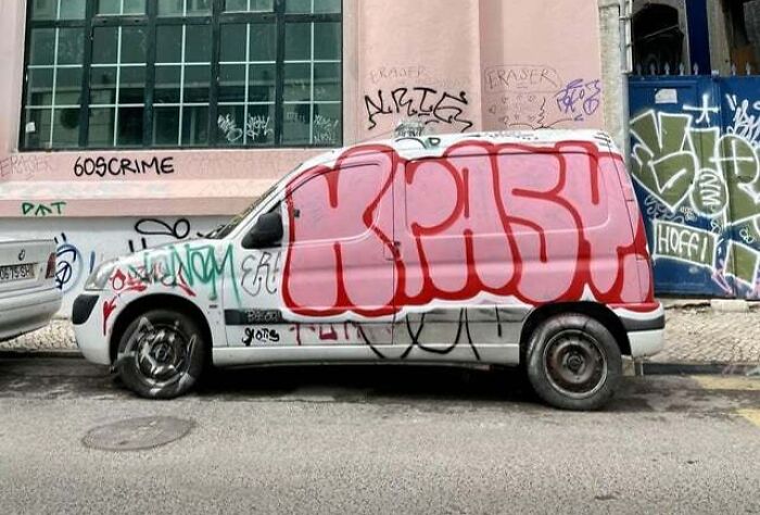 Graffiti art covering a white van with bold pink and red letters, surrounded by tags on urban walls and surfaces.