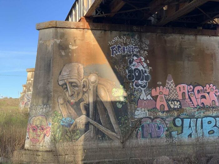 Graffiti art on a concrete bridge wall featuring a detailed elderly figure and colorful abstract designs in urban surroundings.