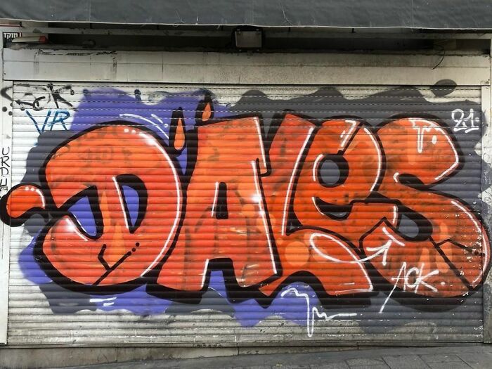Orange graffiti art spelling "Daves" on a metal shutter, showcasing creative graffiti art against a blank urban wall.
