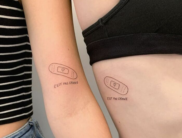 Matching tattoos on arms with text "C'est pas grave" beneath a heart inside a bandage design, embodying word tattoo art.