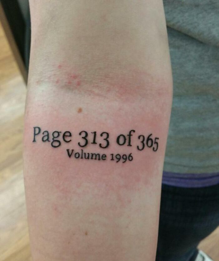 Text tattoo on forearm reads "Page 313 of 365, Volume 1996" in black ink, highlighting word tattoos.
