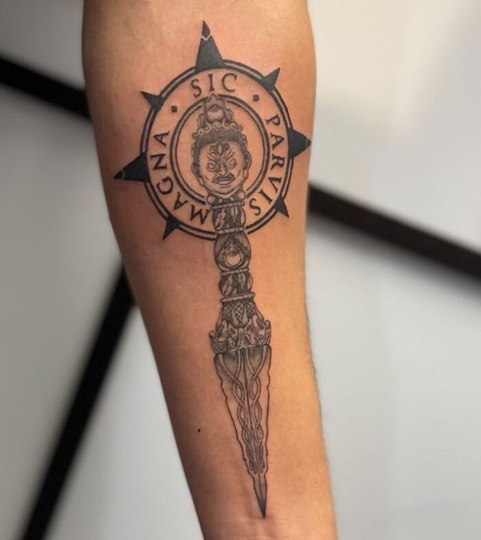 Ornate dagger tattoo on arm with Latin quote, symbolizing deep meaning and connection between heart and skin.