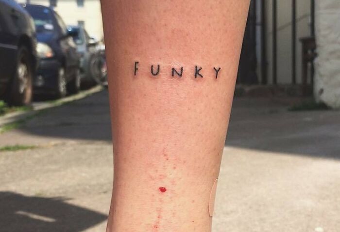 "Word tattoo 'FUNKY' on a person's leg, small and minimalist, set in an outdoor background."
