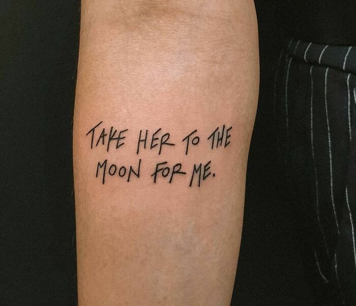 Word tattoo on arm reading "Take her to the moon for me" in black ink.