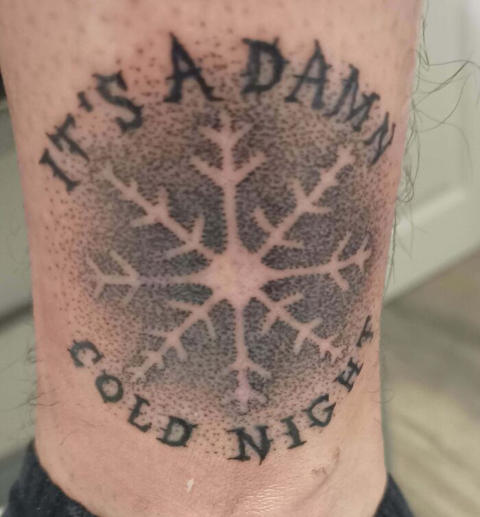 Tattoo of a snowflake with the words "It's a d**n cold night," featuring intricate dotwork and bold lettering.