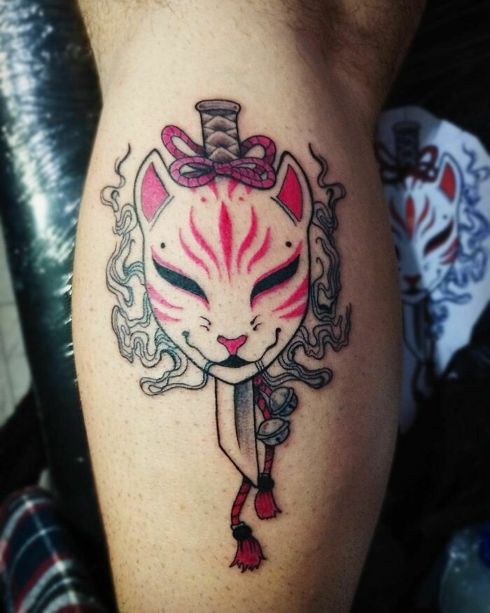 Red ink tattoo of a fox mask with a dagger and decorative elements on a person's calf.