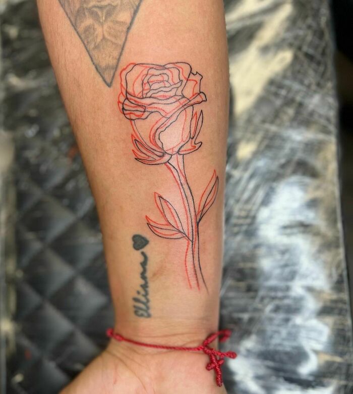 Red ink tattoo of a rose on forearm, with additional fine line details and a name written below.