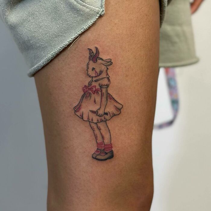 Red ink tattoo of a goat-headed girl in a dress on a person's thigh.